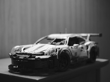 A detailed black and white image of a model race car, likely a LEGO set, displayed on a surface. The model features intricate details such as a rear spoiler, headlamps, and aerodynamic bodywork, creating a realistic impression.