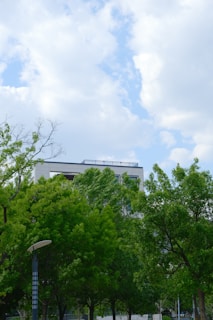 A modern building partially obscured by vibrant green trees under a sky filled with fluffy white clouds. The setting combines urban architecture with natural elements, creating a sense of harmony and tranquility.