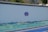A large white wall with a decorative sign in the center. Below the wall is a section covered with a textured, green-blue surface that resembles waves or rolling hills. The ground in front is paved and features two distinct colors: blue and yellow.