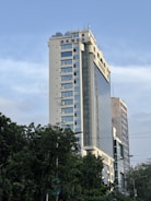 A high-rise building with the letters BKMEA on top, surrounded by other tall buildings and trees at the base. The sky is partly cloudy, and there are some cables visible in the foreground.