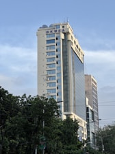 A high-rise building with the letters BKMEA on top, surrounded by other tall buildings and trees at the base. The sky is partly cloudy, and there are some cables visible in the foreground.