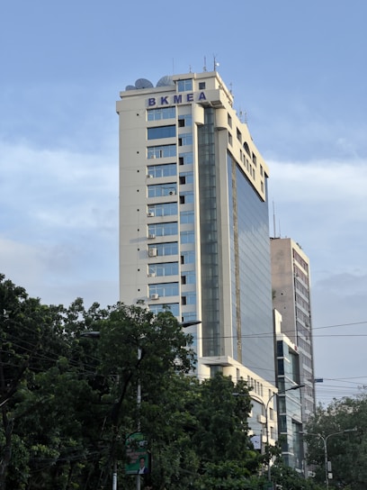 A high-rise building with the letters BKMEA on top, surrounded by other tall buildings and trees at the base. The sky is partly cloudy, and there are some cables visible in the foreground.