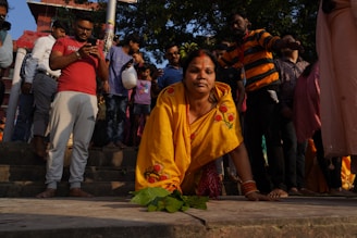 A woman in a yellow sari with floral designs is kneeling on the stairs, surrounded by a crowd of people of various ages. Some individuals are standing and using their phones, while others are observing the scene. The environment appears to be outdoors with trees and a building visible in the background.