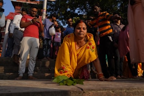 A woman in a yellow sari with floral designs is kneeling on the stairs, surrounded by a crowd of people of various ages. Some individuals are standing and using their phones, while others are observing the scene. The environment appears to be outdoors with trees and a building visible in the background.