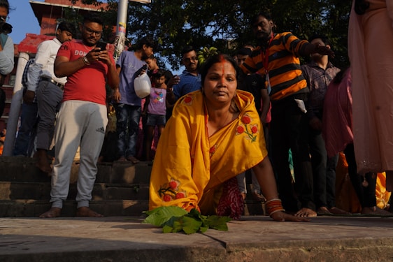 A woman in a yellow sari with floral designs is kneeling on the stairs, surrounded by a crowd of people of various ages. Some individuals are standing and using their phones, while others are observing the scene. The environment appears to be outdoors with trees and a building visible in the background.