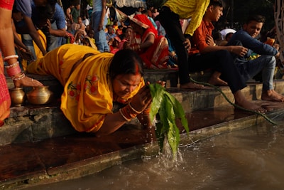 A group of people is gathered near a river. A woman dressed in a yellow saree is bending down to rinse green leaves in the river water. She is surrounded by other people wearing colorful traditional clothing. There are steps leading down to the water, with some people sitting on the steps. Bowls are located near the woman.