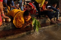 A group of people is gathered near a river. A woman dressed in a yellow saree is bending down to rinse green leaves in the river water. She is surrounded by other people wearing colorful traditional clothing. There are steps leading down to the water, with some people sitting on the steps. Bowls are located near the woman.