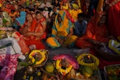 A group of people, primarily women, are seated on the ground, participating in a cultural or religious gathering. They are adorned in vibrant traditional clothing, including sarees in colors such as red, yellow, and blue. In front of them are offerings arranged on the ground, which include fruits, flowers, and decorative items. The setting is outdoors, with many people in the background, suggesting a communal celebration.