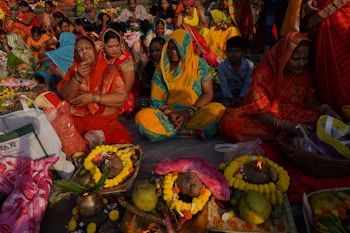 A group of people, primarily women, are seated on the ground, participating in a cultural or religious gathering. They are adorned in vibrant traditional clothing, including sarees in colors such as red, yellow, and blue. In front of them are offerings arranged on the ground, which include fruits, flowers, and decorative items. The setting is outdoors, with many people in the background, suggesting a communal celebration.
