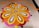 A beautiful and intricate floral design on a tiled floor, created with vibrant petals arranged in concentric patterns. The design features a central circular motif surrounded by petal shapes in a harmonious blend of orange, yellow, pink, red, and white colors. Additional patterns in orange petals form a decorative border.