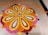 A beautiful and intricate floral design on a tiled floor, created with vibrant petals arranged in concentric patterns. The design features a central circular motif surrounded by petal shapes in a harmonious blend of orange, yellow, pink, red, and white colors. Additional patterns in orange petals form a decorative border.