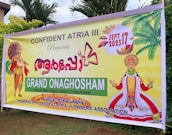 A colorful banner promoting an event called Grand Onaghosham, featuring traditional Kerala imagery including a Kathakali dancer and a mythological figure with an umbrella. The event is set for September 17, 2023, and includes cultural programs. The background includes palm trees and decorative elements.