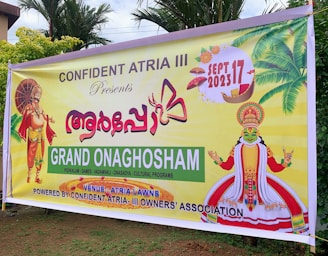 A colorful banner promoting an event called Grand Onaghosham, featuring traditional Kerala imagery including a Kathakali dancer and a mythological figure with an umbrella. The event is set for September 17, 2023, and includes cultural programs. The background includes palm trees and decorative elements.