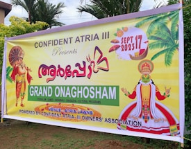 A colorful banner promoting an event called Grand Onaghosham, featuring traditional Kerala imagery including a Kathakali dancer and a mythological figure with an umbrella. The event is set for September 17, 2023, and includes cultural programs. The background includes palm trees and decorative elements.