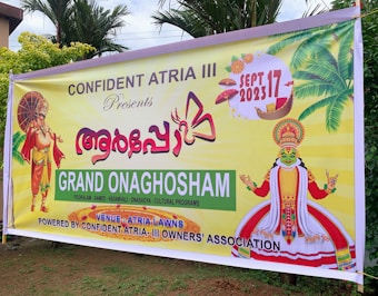 A colorful banner promoting an event called Grand Onaghosham, featuring traditional Kerala imagery including a Kathakali dancer and a mythological figure with an umbrella. The event is set for September 17, 2023, and includes cultural programs. The background includes palm trees and decorative elements.