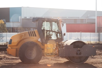 A yellow construction vehicle, specifically a road roller, is parked on a dirt surface in front of a large industrial building. The roller has large, smooth drums for compacting surfaces. Sunlight is creating lens flare effects in the image.