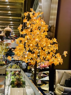 A decorative indoor tree adorned with bright orange leaves and small white lights, set in a modern interior space. The background features metallic decor, acorn-shaped ornaments, and luxurious seating, creating a warm and inviting ambiance.