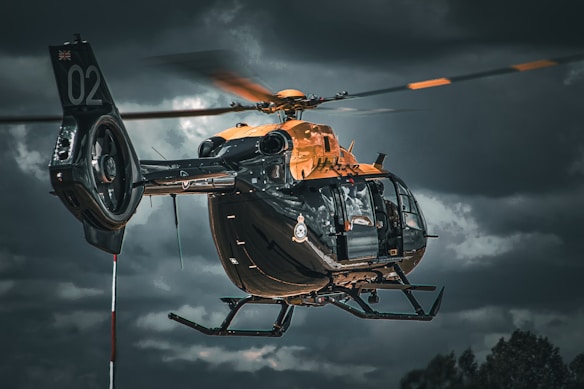 A helicopter is flying against a dramatic cloudy sky backdrop. The helicopter has a sleek design with a primarily dark body accented by orange elements. The image conveys a sense of motion with the rotors blurred in action.