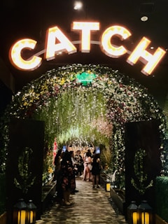 A warmly lit archway adorned with flowers and greenery leads to a lively, crowded area. The word 'CATCH' is prominently displayed in illuminated letters above the arch. Lanterns are placed at the base of the arch, and people dressed up are gathered and socializing inside.