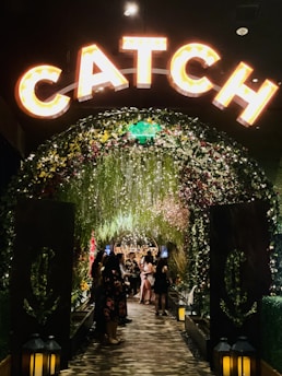 A warmly lit archway adorned with flowers and greenery leads to a lively, crowded area. The word 'CATCH' is prominently displayed in illuminated letters above the arch. Lanterns are placed at the base of the arch, and people dressed up are gathered and socializing inside.
