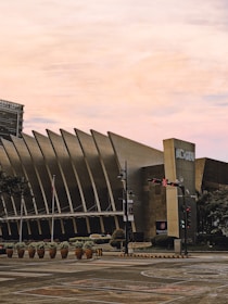 A modern architectural building with a unique design featuring vertical slats and a prominent corner pillar labeled 'ICON'. The scene includes a street with a traffic light showing red, decorative plants in large pots, and street signs. The sky has a warm, soft gradient indicating either sunrise or sunset.