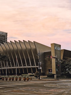 A modern architectural building with a unique design featuring vertical slats and a prominent corner pillar labeled 'ICON'. The scene includes a street with a traffic light showing red, decorative plants in large pots, and street signs. The sky has a warm, soft gradient indicating either sunrise or sunset.
