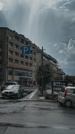 A parking area is adjacent to a multi-story building under an overcast sky. Two cars, one black and one silver, are parked near a blue parking sign. There are also road markings and a sign indicating 'Lake Kawaguchiko.' The atmosphere is calm, with no pedestrians in sight.