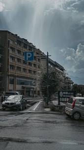 A parking area is adjacent to a multi-story building under an overcast sky. Two cars, one black and one silver, are parked near a blue parking sign. There are also road markings and a sign indicating 'Lake Kawaguchiko.' The atmosphere is calm, with no pedestrians in sight.