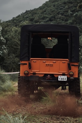 An orange off-road vehicle is driving on a muddy path surrounded by lush greenery and hills. Two people are seated in the jeep, and the back of the jeep is partially covered with a black top. The surrounding landscape is composed of dense foliage and a backdrop of a green hill.