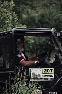 A person with a white beard, wearing a black and red shirt, is seated in an off-road vehicle surrounded by lush green foliage. The vehicle has the number 207 displayed prominently along with various stickers related to an off-road challenge event.