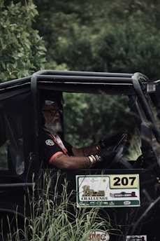 A person with a white beard, wearing a black and red shirt, is seated in an off-road vehicle surrounded by lush green foliage. The vehicle has the number 207 displayed prominently along with various stickers related to an off-road challenge event.