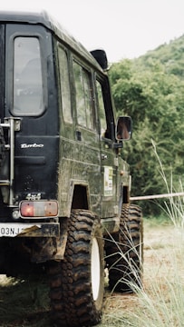 A rugged four-wheel-drive vehicle parked on uneven, grassy terrain surrounded by dense green foliage. The vehicle has large, mud-covered tires and a black, slightly worn exterior.