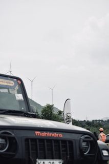 A black Mahindra vehicle is partially visible in the foreground with a logo and stickers on the windshield. In the background, there are several wind turbines against a cloudy sky, with hills and greenery. A person wearing an orange safety vest and helmet stands near a banner.