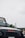 A black Mahindra vehicle is partially visible in the foreground with a logo and stickers on the windshield. In the background, there are several wind turbines against a cloudy sky, with hills and greenery. A person wearing an orange safety vest and helmet stands near a banner.