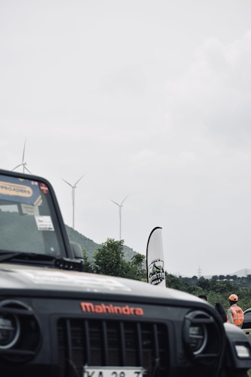 A black Mahindra vehicle is partially visible in the foreground with a logo and stickers on the windshield. In the background, there are several wind turbines against a cloudy sky, with hills and greenery. A person wearing an orange safety vest and helmet stands near a banner.