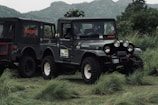 A rugged off-road vehicle parked on lush green grass with hills in the background. The vehicle has a military-style design with multiple round headlights, large tires, and visible branding on its side and back panel.