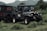 A rugged off-road vehicle parked on lush green grass with hills in the background. The vehicle has a military-style design with multiple round headlights, large tires, and visible branding on its side and back panel.