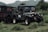 A rugged off-road vehicle parked on lush green grass with hills in the background. The vehicle has a military-style design with multiple round headlights, large tires, and visible branding on its side and back panel.