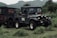 A rugged off-road vehicle parked on lush green grass with hills in the background. The vehicle has a military-style design with multiple round headlights, large tires, and visible branding on its side and back panel.