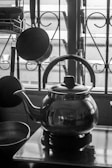 A black and white photograph of a kitchen setup featuring a metallic kettle prominently placed on a stovetop. Behind the kettle, several kitchen utensils such as a hanging ladle and spoon are visible, alongside a window with wrought iron bars. The overall scene is lit from behind, creating reflections on the shiny surface of the kettle.