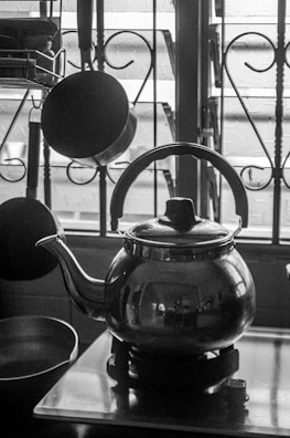A black and white photograph of a kitchen setup featuring a metallic kettle prominently placed on a stovetop. Behind the kettle, several kitchen utensils such as a hanging ladle and spoon are visible, alongside a window with wrought iron bars. The overall scene is lit from behind, creating reflections on the shiny surface of the kettle.