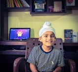 A young child wearing a white turban and a striped navy shirt sits on a chair, smiling. Behind him is a desk with a laptop displaying a colorful abstract wallpaper. Shelves with books and other items are mounted on the wall above the desk.