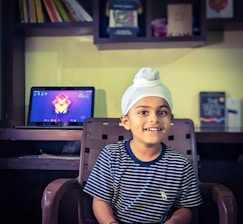 A young child wearing a white turban and a striped navy shirt sits on a chair, smiling. Behind him is a desk with a laptop displaying a colorful abstract wallpaper. Shelves with books and other items are mounted on the wall above the desk.