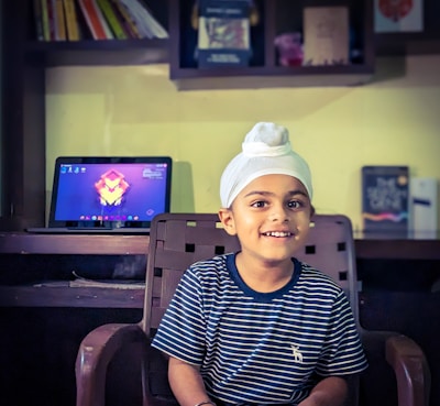 A young child wearing a white turban and a striped navy shirt sits on a chair, smiling. Behind him is a desk with a laptop displaying a colorful abstract wallpaper. Shelves with books and other items are mounted on the wall above the desk.