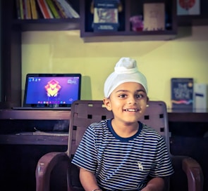 A young child wearing a white turban and a striped navy shirt sits on a chair, smiling. Behind him is a desk with a laptop displaying a colorful abstract wallpaper. Shelves with books and other items are mounted on the wall above the desk.