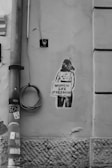 A graffiti artwork on a textured, cracked wall depicts a silhouette of a person holding a sign that reads 'Women Life Freedom.' The wall has an old, worn appearance, with a vertical pipe and coiled cable mounted on it. Black and white stickers are attached to the pipe.