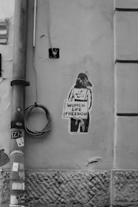 A graffiti artwork on a textured, cracked wall depicts a silhouette of a person holding a sign that reads 'Women Life Freedom.' The wall has an old, worn appearance, with a vertical pipe and coiled cable mounted on it. Black and white stickers are attached to the pipe.