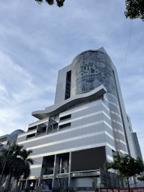 A modern, multi-story building with a sleek, metallic design. The facade features a large cylindrical structure with signage for a theme park. Glass windows and geometric patterns adorn the exterior, while palm trees add a touch of greenery at ground level.