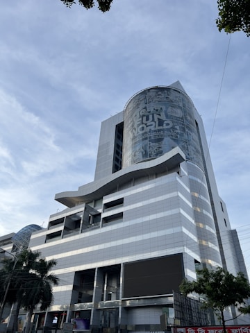 A modern, multi-story building with a sleek, metallic design. The facade features a large cylindrical structure with signage for a theme park. Glass windows and geometric patterns adorn the exterior, while palm trees add a touch of greenery at ground level.