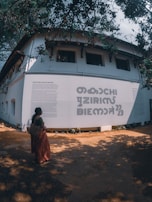 A person in traditional attire stands in front of a large white wall with bold text and smaller text paragraphs. The wall is part of a building with multiple windows, and the scene is shaded by overhanging tree branches and leaves.